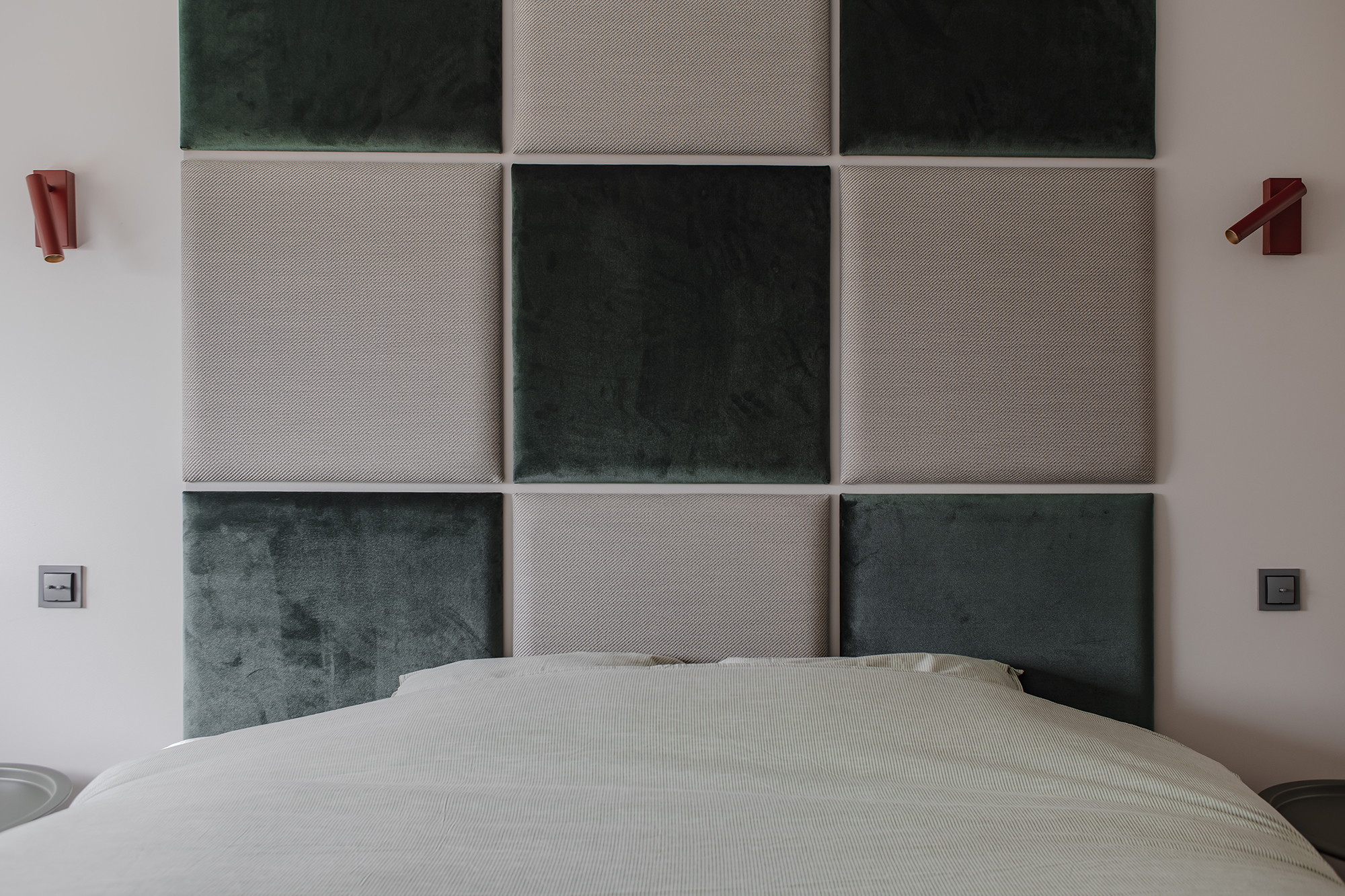 Zabawa formą, teksturą i kolorem, czyli tapicerowane różnymi tkaninami panele ścienne, ułożone w szachownicę i podkreślone czerwonymi kinkietami.